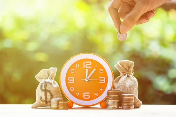 Money savings, Investment, time and money growing concept : Stacking growing coins, Moneybags and orange clock on wooden table. Saves money for the future. Time investment use money for work.