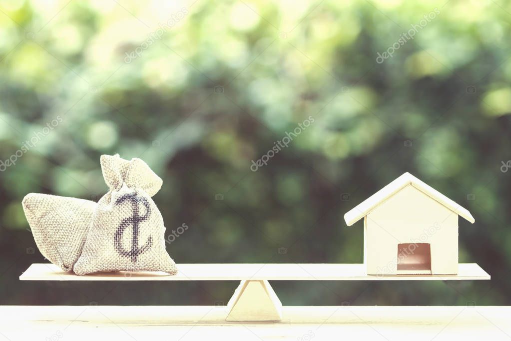 Home loan, mortgages, debt, savings money for home buying concept : Balance a money bag and small residential, house model on table against green nature background. Exchange of finances and houses.