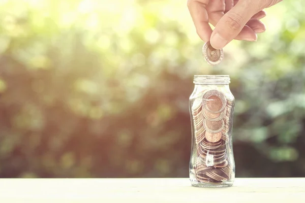 Financial investment / Savings money growing concept : Hand holding coin over coins in glass jar on wood table with nature and space. Conceptual saving money for growing business and life future.