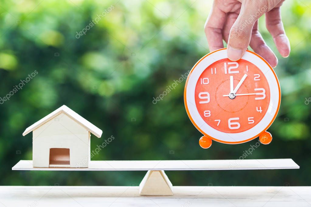 Home bargain, real estate, loan, house lender reverse mortgage concept. Balance orange clock and hand holding small residence on wooden table and blurred green nature as background. Time and home.