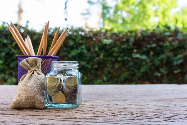 Money savings, investment, making money for future, financial wealth management concept. Coins in glass jar and pencil. Cash in hemp bags or burlap sacks on wood tables. Green nature background.