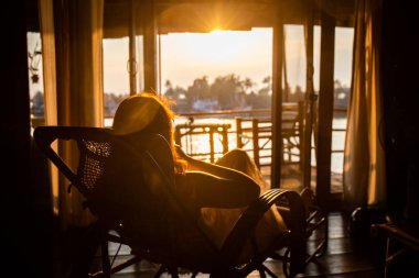Samut Songkhram, Tayland 'da altın saat gün batımında sallanan sandalyede oturan kadın, ahşap bir evde güneşe bakıyor.