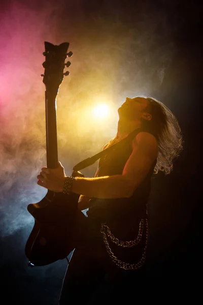 Young man with long hair with electric guitar in neon lights. Rock musician plays in atmosphere of stage