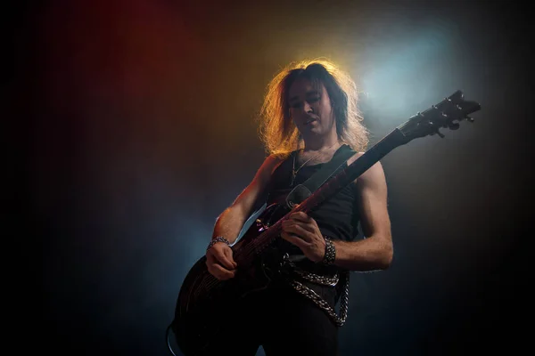 Young man with long hair with electric guitar in neon lights. Rock musician plays in atmosphere of stage