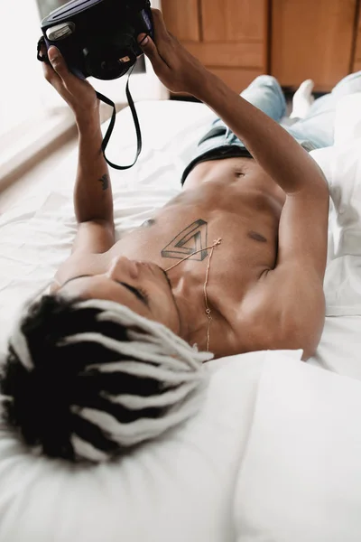 Portrait of handsome african black man with dreadlocks lying with snapshot camera and doing selfie in bed at home. Contrast black and white — Stock Photo, Image