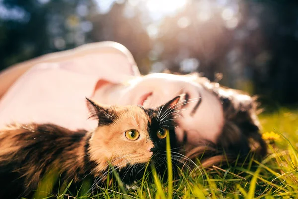 Chica acostada en la hierba con el gato. Concepto de clima cálido de primavera o verano. Fondo Bokeh. Gatito de jengibre con máscara de dos colores —  Fotos de Stock