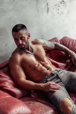 Moda portre dövme ve sıcak poz kırmızı deri kanepede oturan siyah sakalı ile seksi çıplak erkek modeli. Çatı katı odası iç gri beton duvar ile. Profesyonel Studio resim.