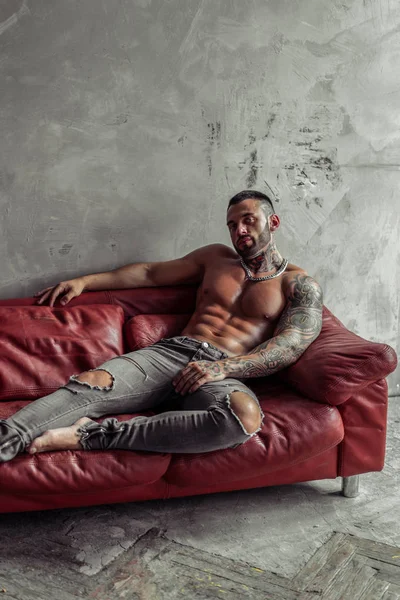 Moda portre dövme ve sıcak poz kırmızı deri kanepede oturan siyah sakalı ile seksi çıplak erkek modeli. Çatı katı odası iç gri beton duvar ile. Profesyonel Studio resim.