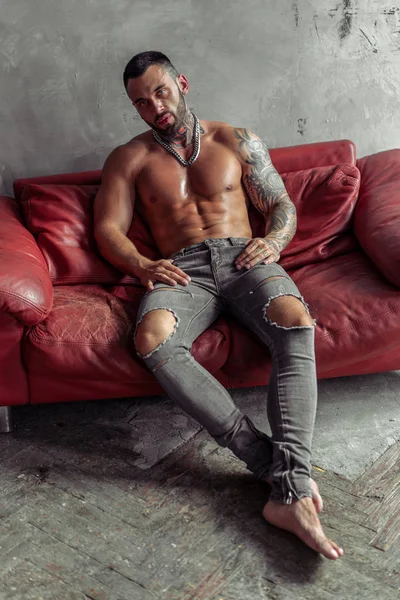 Moda portre dövme ve sıcak poz kırmızı deri kanepede oturan siyah sakalı ile seksi çıplak erkek modeli. Çatı katı odası iç gri beton duvar ile. Profesyonel Studio resim.