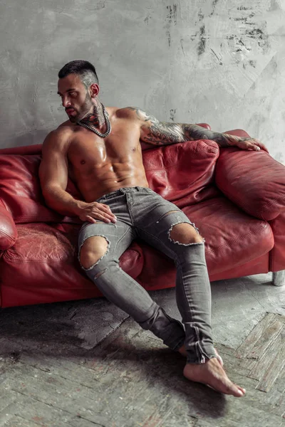 Moda portre dövme ve sıcak poz kırmızı deri kanepede oturan siyah sakalı ile seksi çıplak erkek modeli. Çatı katı odası iç gri beton duvar ile. Profesyonel Studio resim.