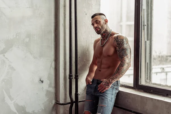Moda portre dövme ve siyah sakallı ayakta pencerenin yakınındaki üzerinde sıcak poz ile seksi çıplak erkek modeli. Çatı katı odası iç gri beton duvar ile. Profesyonel Studio resim.