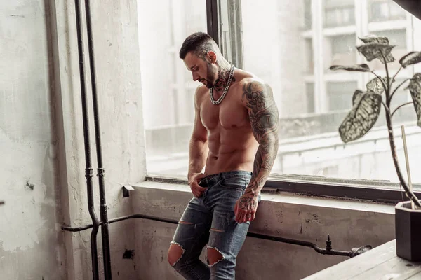 Moda portre dövme ve siyah sakallı ayakta pencerenin yakınındaki üzerinde sıcak poz ile seksi çıplak erkek modeli. Çatı katı odası iç gri beton duvar ile. Profesyonel Studio resim.