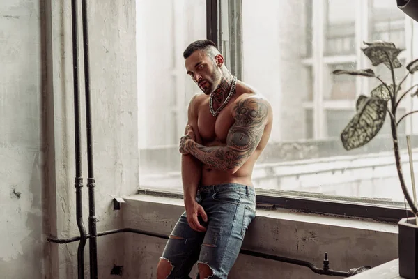 Moda portre dövme ve siyah sakallı ayakta pencerenin yakınındaki üzerinde sıcak poz ile seksi çıplak erkek modeli. Çatı katı odası iç gri beton duvar ile. Profesyonel Studio resim.