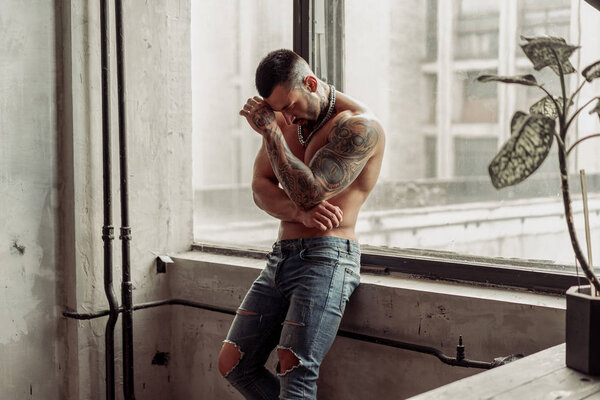 Fashion portrait of Sexy naked male model with tattoo and a black beard standing in hot pose on near the window. Loft room interior with grey concrete wall. Professional Studio image.