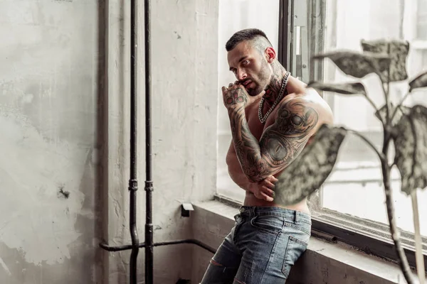 Seksi çıplak erkek modeli dövme ve magic yakınındaki penceresi içinde sıcak poz ayakta gözleri portresi kapatın. Çatı katı odası iç gri beton duvar ile. Profesyonel Studio resim.