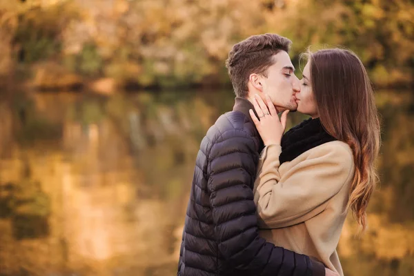 Couple in love standing in autumn park with yellow fallen leaves. Man and woman enjoying a day together. Boy kissing a girl in lips