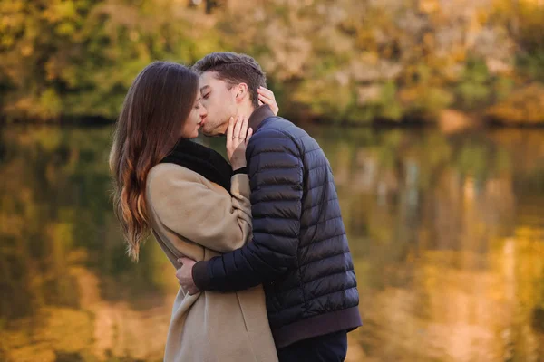 Couple in love standing in autumn park with yellow fallen leaves. Man and woman enjoying a day together. Boy kissing a girl in lips