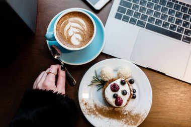 Üstten Görünüm tasarımcıları yer. Kadın el tutmak kaşık. Serbest çalışma alanı. Defter ile cep telefonu, fincan kahve ve tatlı ahşap tablo