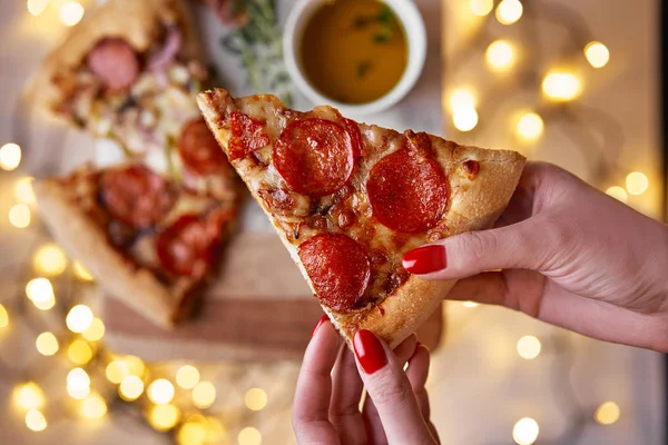 Noel ve yeni yıl atmosfer. Kadının el dilim domates, biber ve beyaz mermer kesme tahtası üzerinde peynir erime ile İtalyan pizza alır. Arka plan ile Işıklar bokeh ve seçici