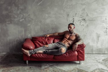 Dövme ve kırmızı deri kanepe üzerinde sıcak poz yalan siyah sakallı Seksi çıplak erkek modeli Moda portresi. Gri beton duvarile Loft oda iç. Profesyonel Stüdyo görüntüsü