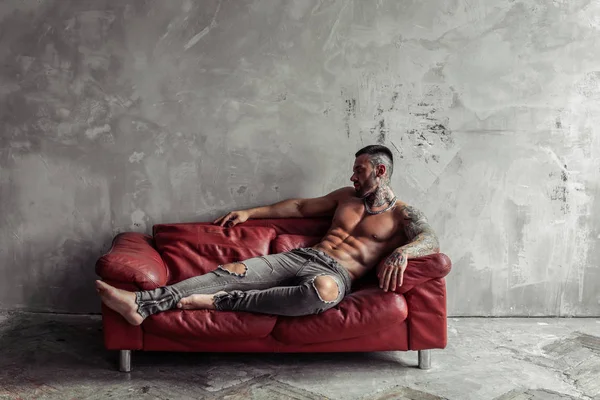 Dövme ve kırmızı deri kanepe üzerinde sıcak poz yalan siyah sakallı Seksi çıplak erkek modeli Moda portresi. Gri beton duvarile Loft oda iç. Profesyonel Stüdyo görüntüsü