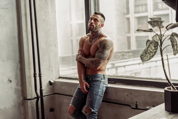 Moda portre dövme ve siyah sakallı ayakta pencerenin yakınındaki üzerinde sıcak poz ile seksi çıplak erkek modeli. Çatı katı odası iç gri beton duvar ile. Profesyonel Studio resim