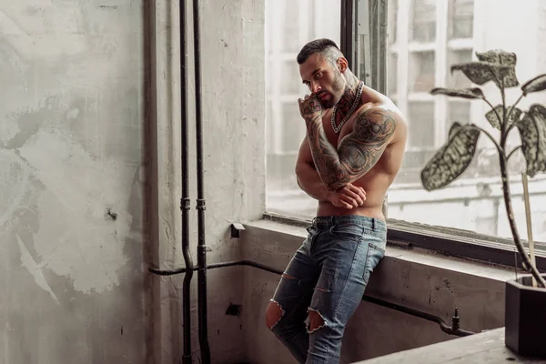 Moda portre dövme ve siyah sakallı ayakta pencerenin yakınındaki üzerinde sıcak poz ile seksi çıplak erkek modeli. Çatı katı odası iç gri beton duvar ile. Profesyonel Studio resim