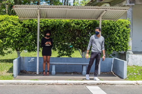 two young people at a bus stop wearing black masks keeping the social distance