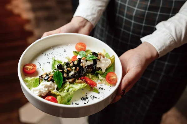 Garson bir tabak beyaz bir tabak tutuyor, yakın plan ellerle. Kiraz domatesli salata, fesleğen ve çam fıstığı.
