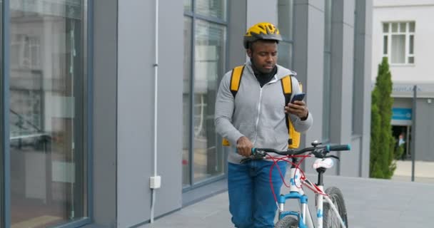 Afro-americano jovem entregador em casco levando sua bicicleta na rua, tocando no celular e procurando endereço. Mensageiro masculino usando navegador e andando com bicicleta. Um tipo a passear . — Vídeo de Stock