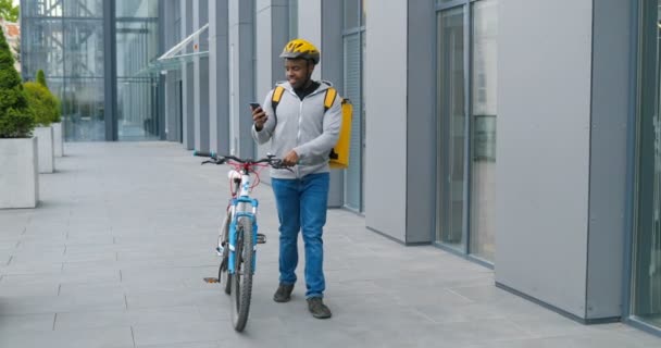 Joven ciclista afroamericano en casque que conduce su bicicleta en la calle, tocando y enviando mensajes de texto en el teléfono inteligente. Mensajero hombre utilizando el teléfono móvil y caminar con bicicleta. Un tipo dando un paseo . — Vídeos de Stock