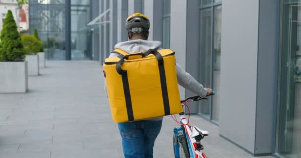 Traseira de correio masculino afro-americano com saco grande de volta em casco andando e levando bicicleta na rua e entregando alimentos ou parcelas. Visão traseira no ciclista entregador ter uma caminhada com bicicleta . — Vídeo de Stock