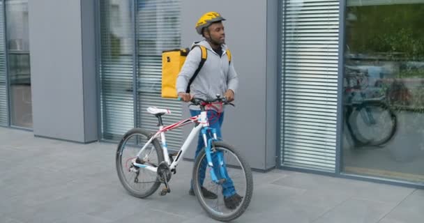 Joven guapo afroamericano en casco caminando y liderando la bicicleta mientras trabajaba como repartidor. Entrega del concepto de bicicleta durante el bloqueo . — Vídeos de Stock