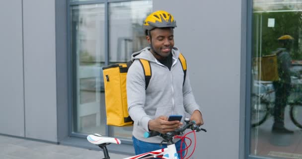 Afro-americano jovem entregador em casco em pé em sua bicicleta na rua, tocando e mensagem de texto no smartphone. À procura de morada. mensageiro masculino usando a navegação no telefone para encontrar endereço . — Vídeo de Stock