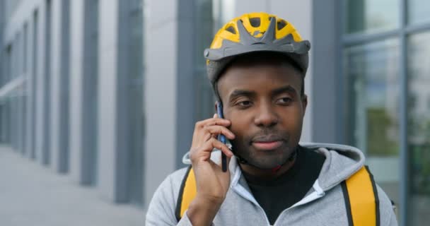 Africano americano jovem bonito ciclista masculino em casco falando no telefone celular ao ar livre. Entregador com mochila falando no celular na rua . — Vídeo de Stock