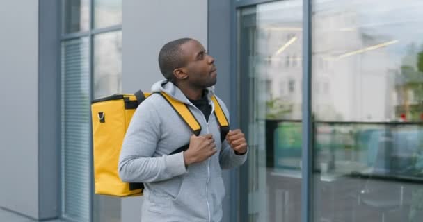 Afrikai-amerikai jóképű fiatalember sétál az utcán nagy sárga hátizsákkal, miközben futárként dolgozik. A koncepció megvalósítása gyalogosan. — Stock videók