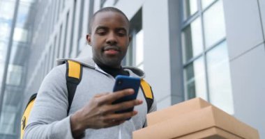 Elinde karton paketlerle caddede bekleyen Afro-Amerikalı teslimatçı. Adres bulmak için telefon üzerinden navigasyon haritasını dinliyor. Erkek kurye, kutuları teslim etmek için akıllı telefonda navigasyon sistemini kullanıyor..