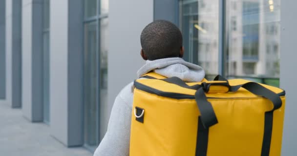 Posteriore di corriere afroamericano maschio con grande borsa sul retro passeggiando per strada e consegnando cibo o pacchi. Indietro vista sul fattorino alla ricerca di indirizzo per consegnare . — Video Stock