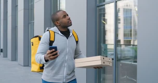 Afro-Amerikaanse jonge bezorger loopt op straat, tikt op mobiele telefoon en scrollt terwijl hij op zoek is naar adres. Mannelijke koerier met pizza dozen met een wandeling en het gebruik van navigator op de smartphone. — Stockvideo