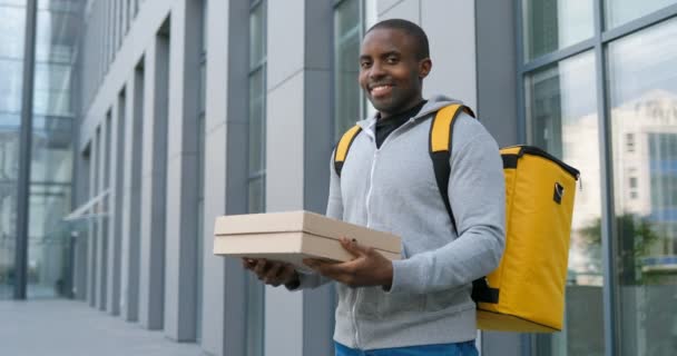 Portrét veselého afroamerického doručovatele stojícího na ulici, držícího kartónové krabice a usmívajícího se. Samec kurýr s pizzou v ruce dívá na kameru s úsměvem. Venku. — Stock video