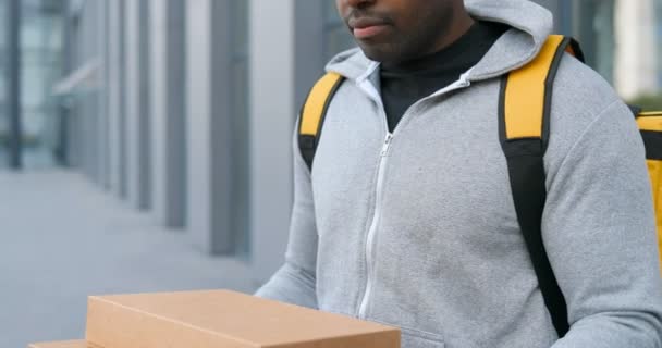 Portrét pohledného afroamerického doručovatele stojícího na ulici a držícího kartónové krabice. Muž kurýr s balíčky dívá na kameru a usmívá se venku. — Stock video