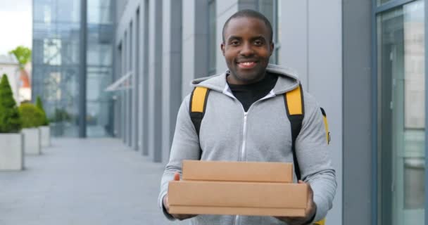 African American young cheerful deliveryman in backpack giving parcels to camera at street. Male courier with bag bringing boxes to customer and handing outdoor. — Stock Video