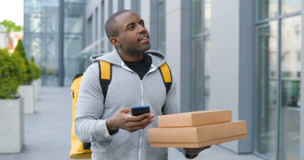 Repartidor afroamericano con paquetes de cartón de pie en la calle, tocando en el mapa de navegación en el teléfono en busca de dirección. Correo masculino utilizando el sistema de navegación en el teléfono inteligente para entregar cajas . — Vídeos de Stock
