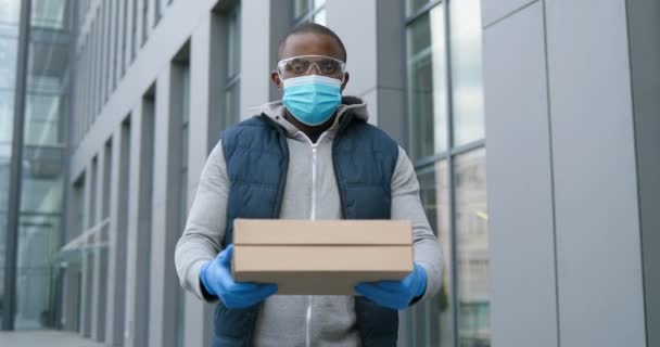 Portretfoto van een knappe Afro-Amerikaanse bezorger in een bril, handschoenen en medisch masker op straat met kartonnen dozen die aan de camera geven. Mannelijke koerier overhandigt pizza aan klant. — Stockvideo
