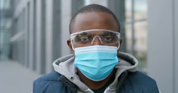 Großaufnahme des Gesichts eines jungen, gut aussehenden Afroamerikaners mit medizinischer Maske und Brille, der direkt in die Kamera schaut. Porträt eines männlichen Kuriers auf der Straße während des Coronavirus. Quarantäne-Konzept. — Stockvideo