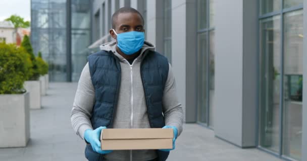 穿着手套和医疗面罩站在街头，拿着并递给纸盒的英俊非洲裔美国人的肖像照片。男信使送披萨和看相机. — 图库视频影像