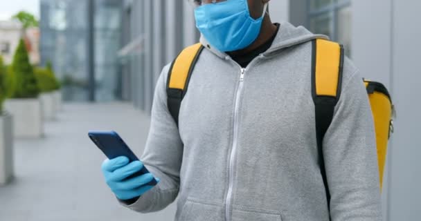 Close up of African American man in medical mask and gloves outdoor texting message on smartphone. Deliveryman tapping and typing on mobile phone, using map navigator and looking for address. — Stock Video