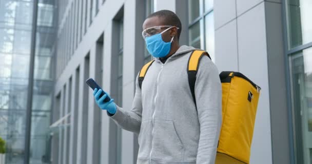 Hombre afroamericano en máscara médica, gafas y guantes mensaje de texto al aire libre en el teléfono inteligente. Entregador tocando y escribiendo en el teléfono móvil, utilizando el navegador de mapas y en busca de dirección . — Vídeo de stock