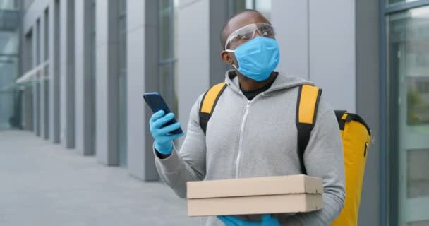 Seorang pria Afrika-Amerika yang membawa kacamata, sarung tangan dan masker medis berdiri di jalan dan memegang kotak pizza sambil mengirim pesan lewat telepon. Kurir pria mengetuk dan menggulung di smartphone. — Stok Video