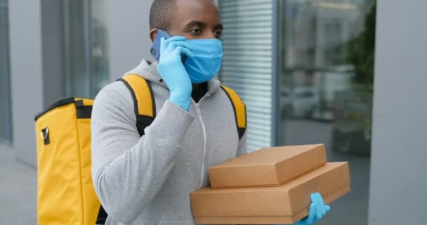 Afro-Amerikaanse jonge bezorger in medisch masker en handschoenen lopen op straat, praten op mobiele telefoon en op zoek naar adres. Man koerier met dozen met een wandeling en spreken op mobiele telefoon. — Stockvideo